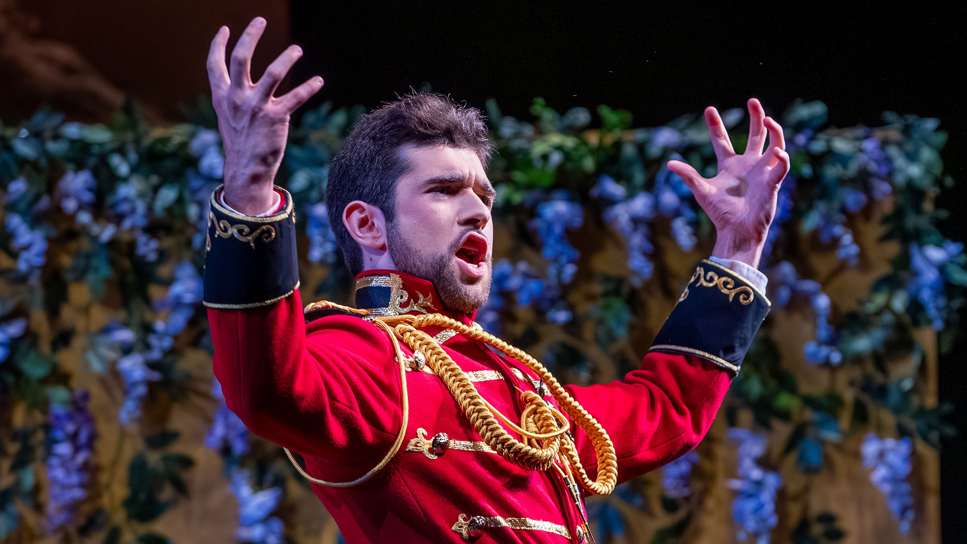 An actor on stage, dressed in a vibrant red, gold-trimmed military-style jacket, singing dramatically with both hands raised, against a backdrop of dark green leaves and purple wisteria. The text THEATRE is in the lower right corner.
