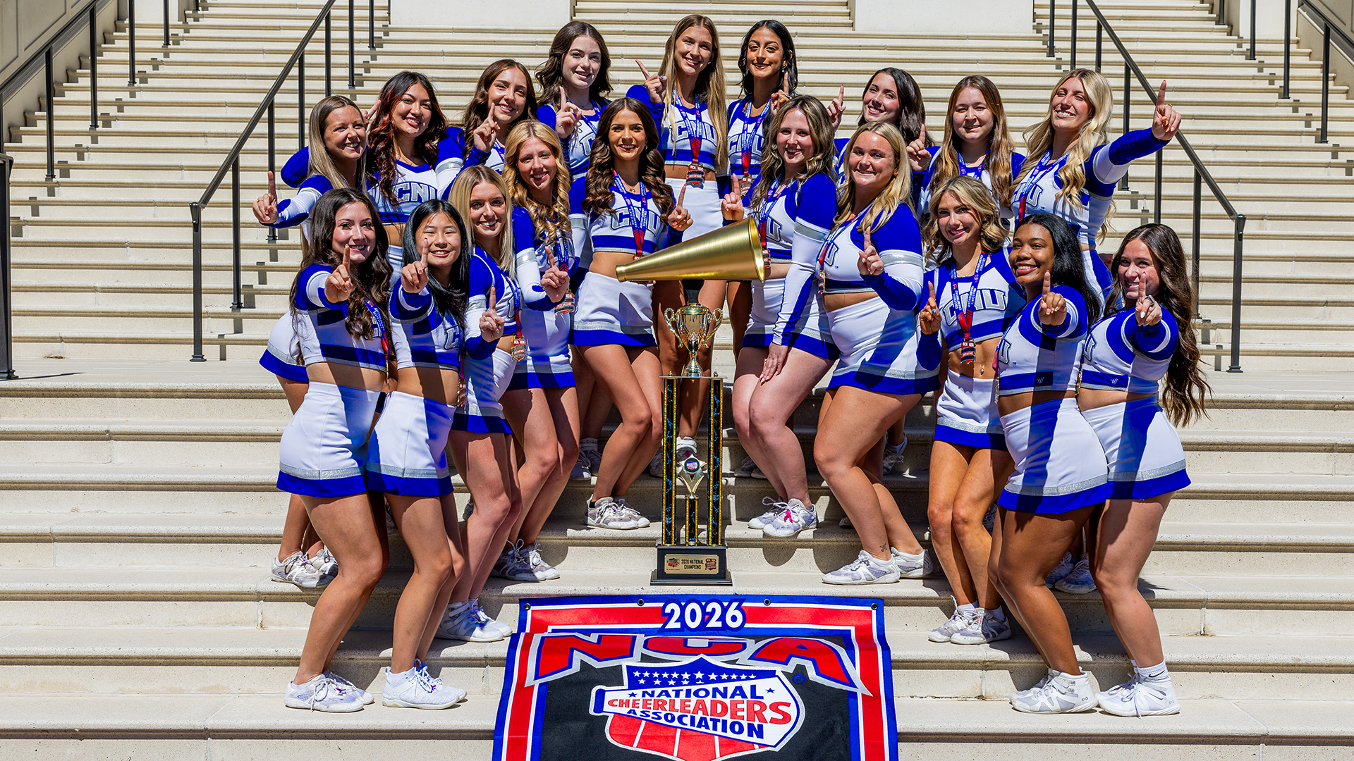 Members of the 2026 National Championship winning cheerleading squad on the steps of Christopher Newport Hall with a large trophy and banner