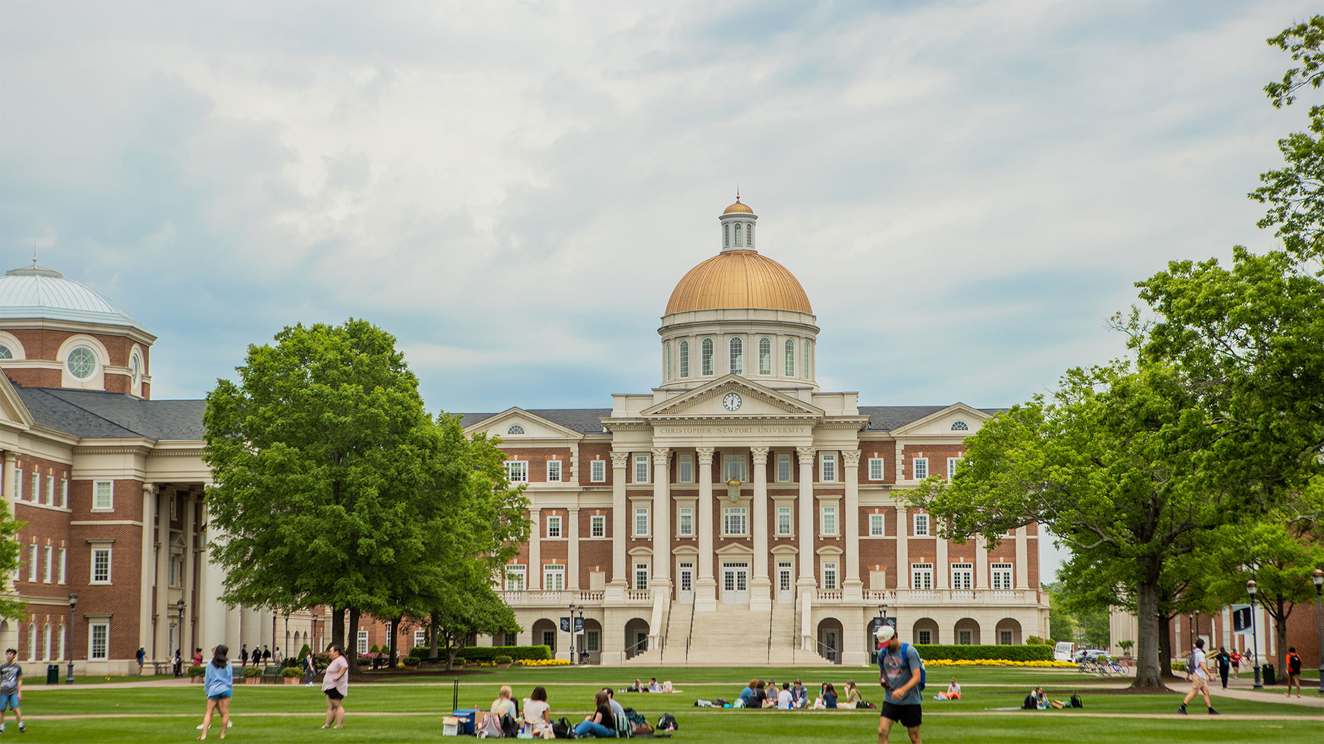 Students enjoying warm weather on the Great Lawn with Christopher Newport Hall in the background against a partly grey sky