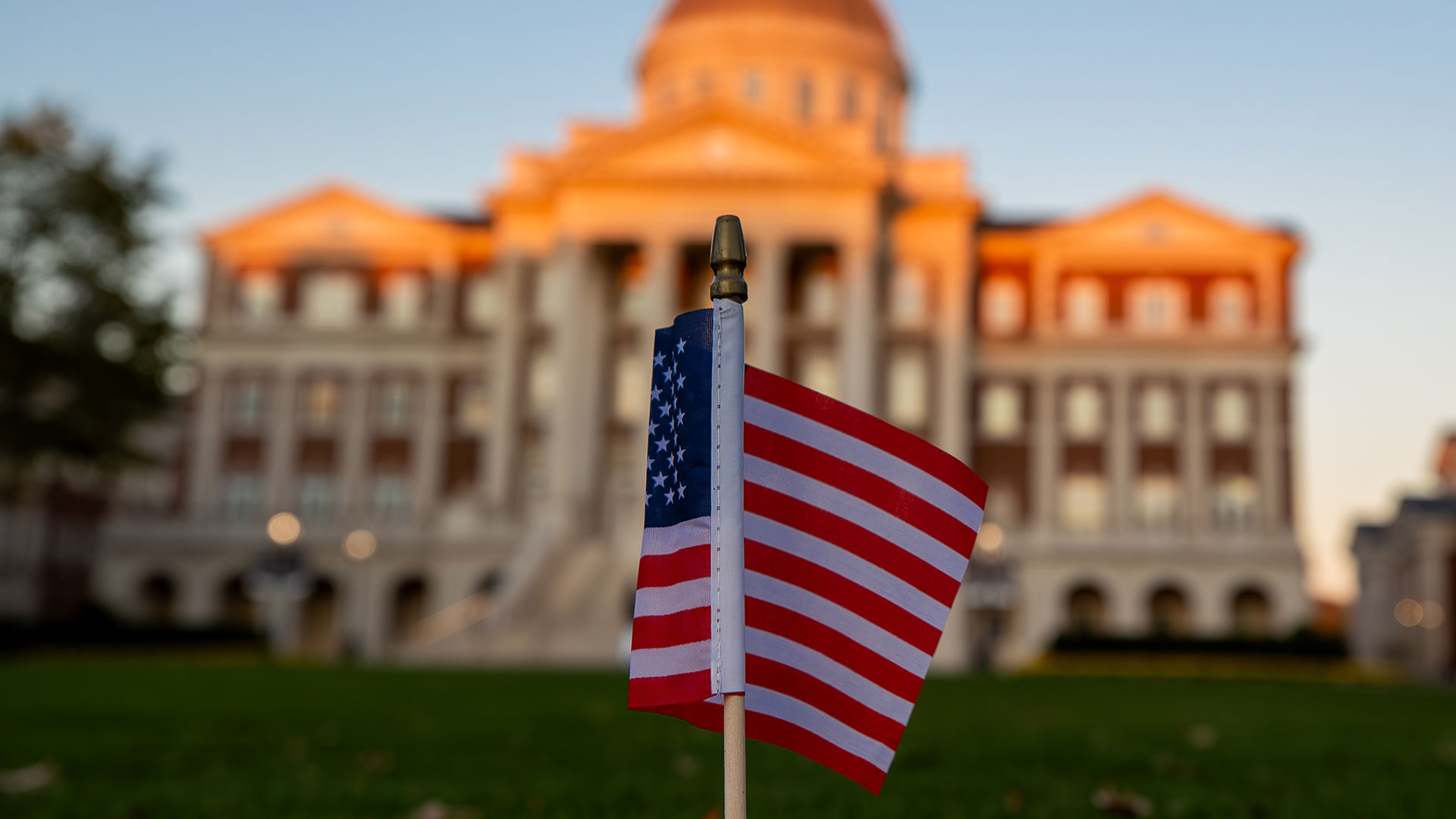 A small American flag is planted in the grass with a large, domed university building softly focused in the background at sunset.