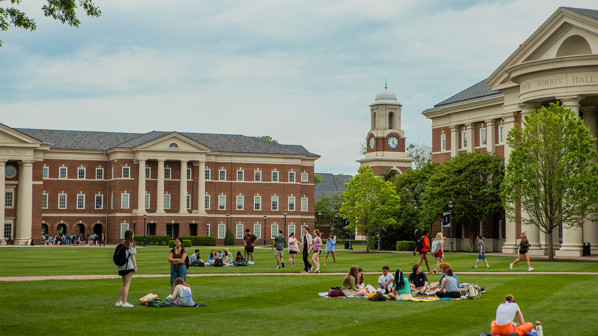 Students sit on the Great Lawn with the McMurran Hall, the Bell Tower and Forbes Hall behind them.