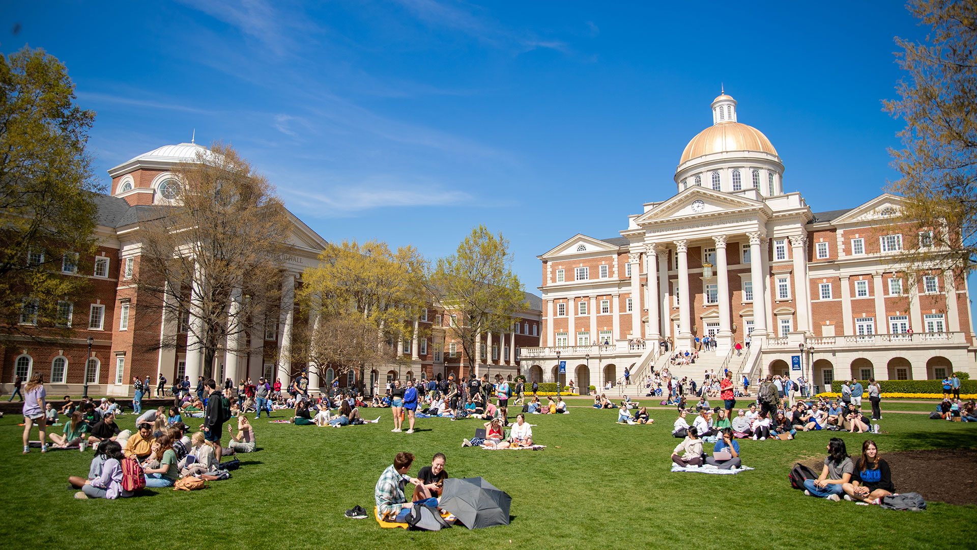 Students sit on blankets in front of Christopher Newport Hall on a sunny day.