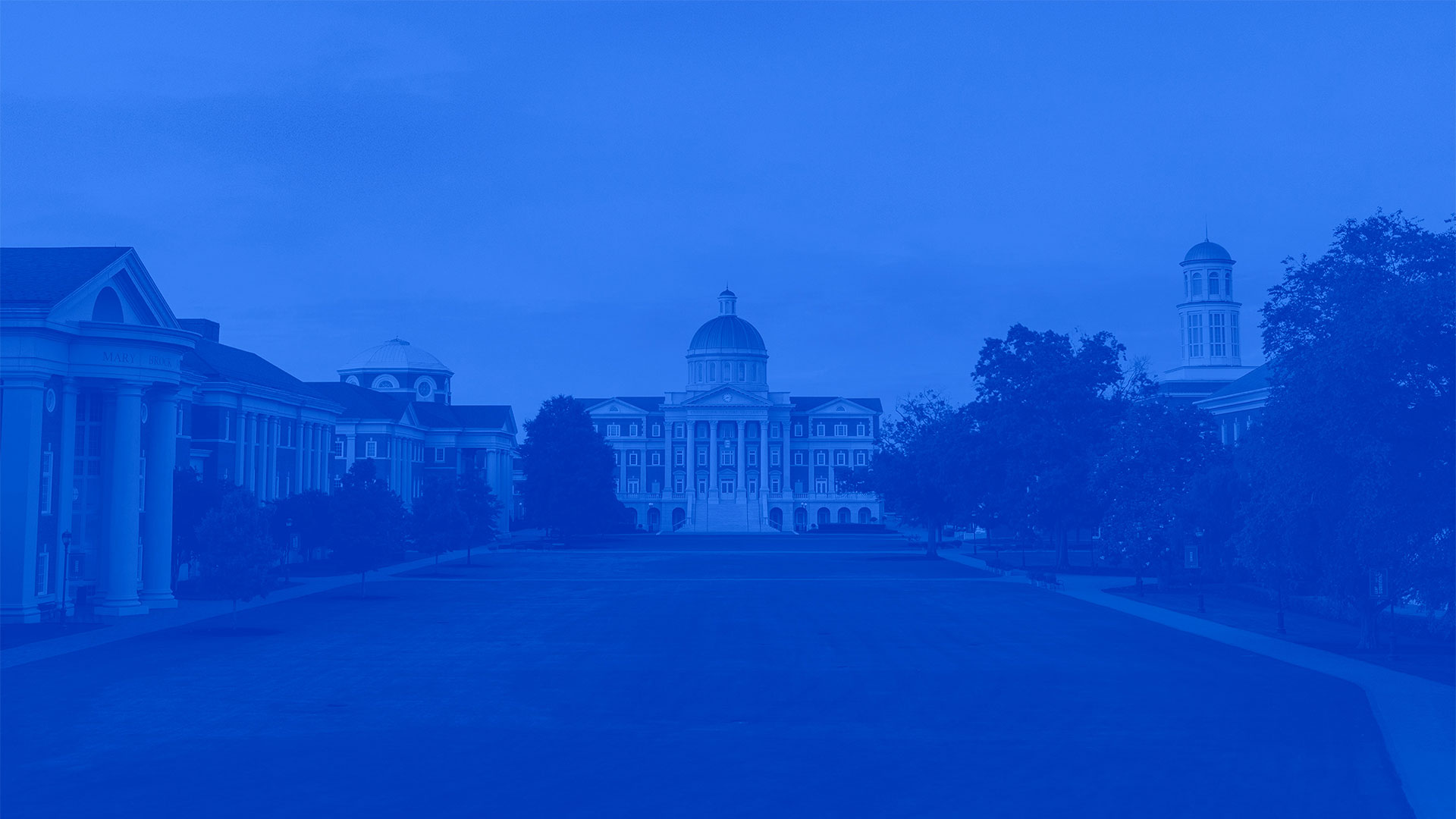 A wide view of the campus of Christopher Newport University at dusk, featuring Christopher Newport Hall with its prominent golden dome and cupola at the head of a large, grassy lawn. Stately academic buildings with classical architecture and columns line both sides of the lawn under a deep blue sky with a blue filter