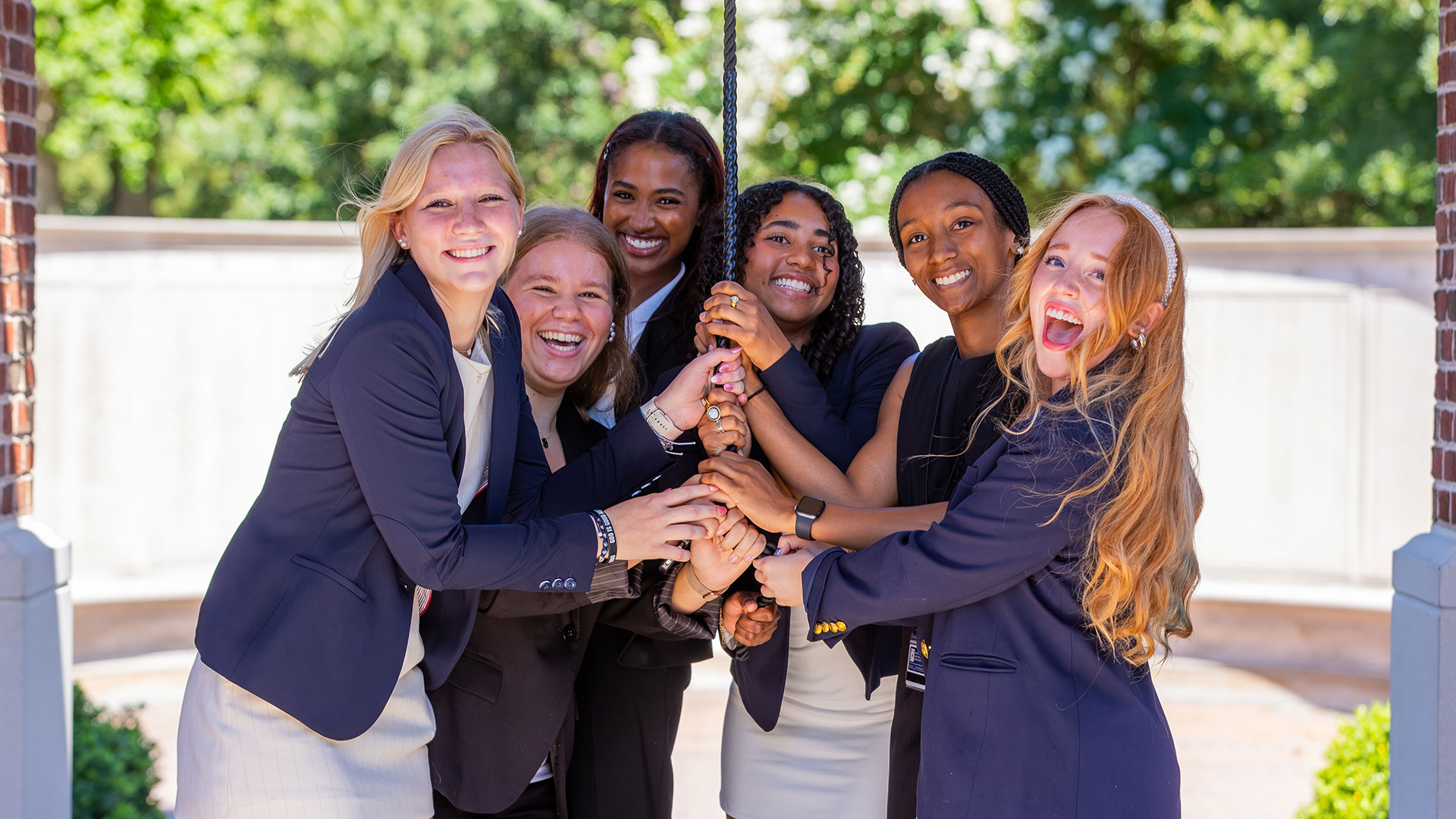 A group of girls in business attire gather and pull on a rope to ring a bell in the bell tower.
