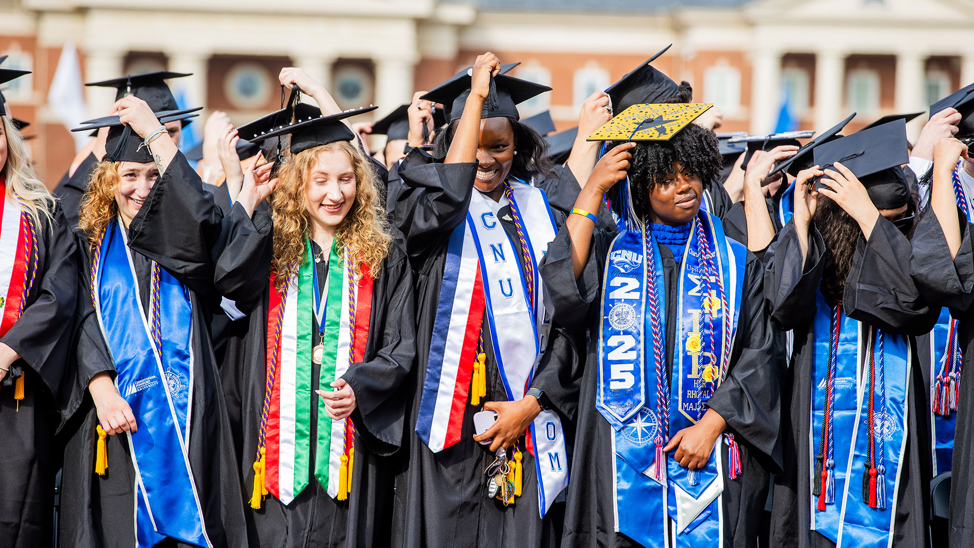 Graduates in black caps and gowns and multi-colored stoles and cords turn the tassel from one side of their cap to another during the conferral ceremony.
