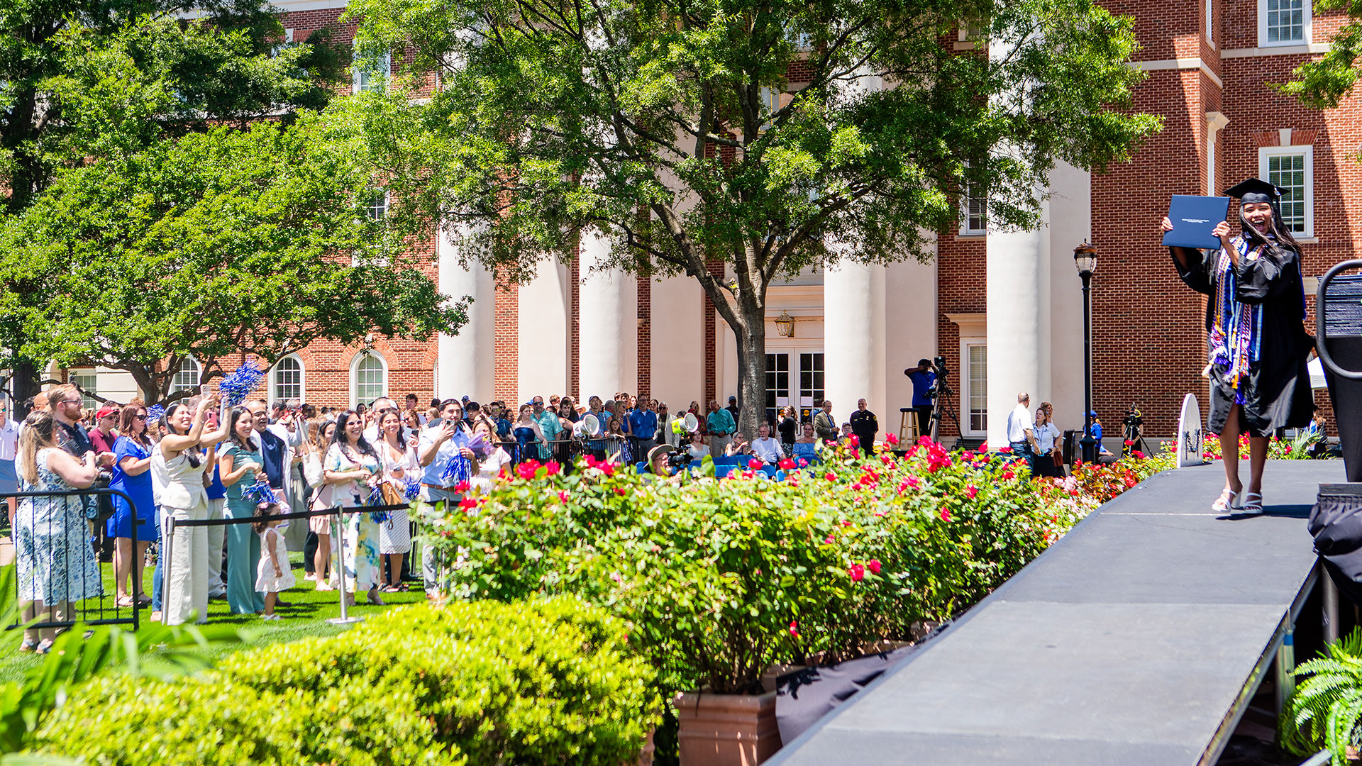 - a joyful graduate holds up her diploma on an outdoor stage while a crowd cheers from the ground.