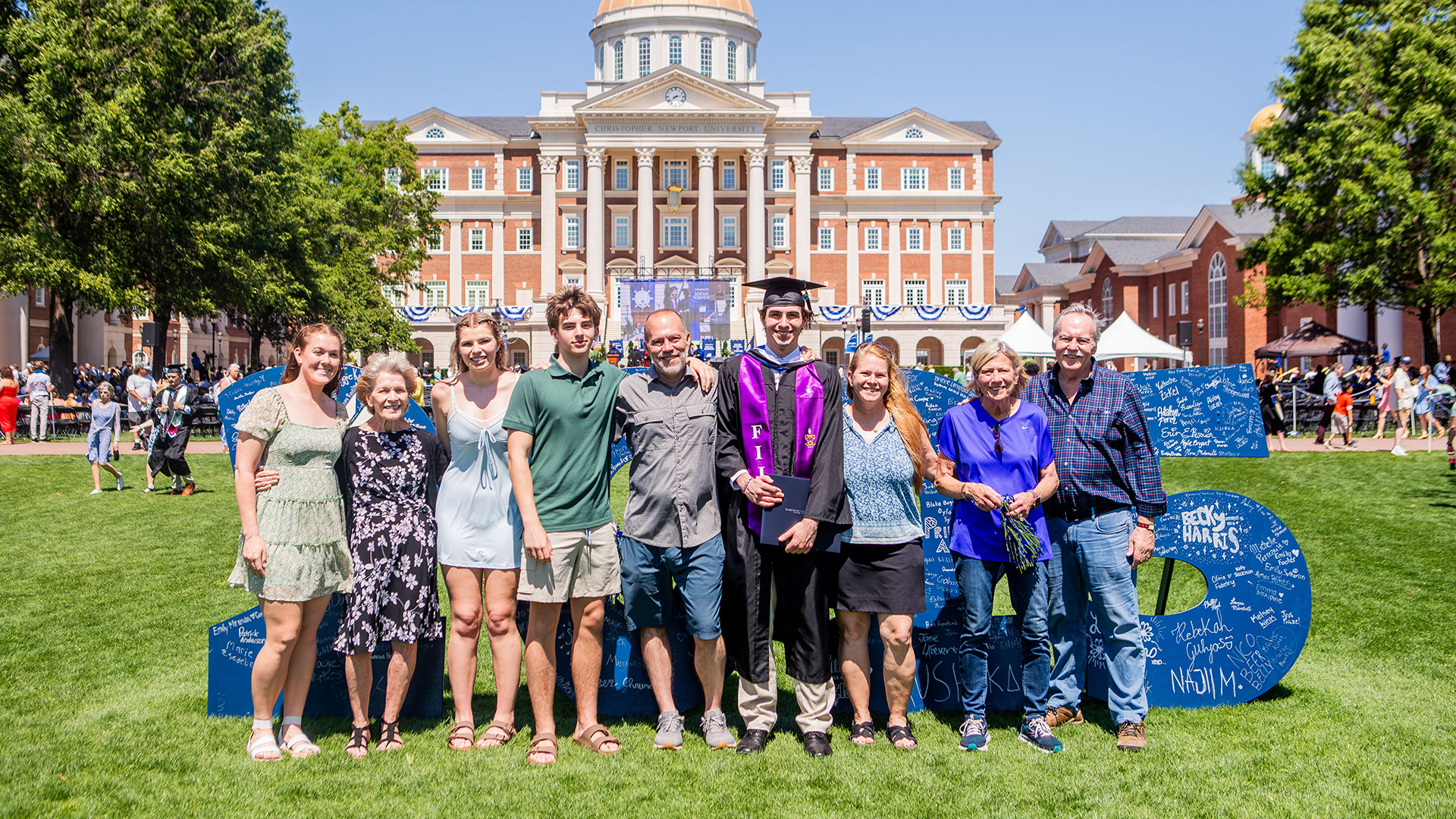 - A graduate in reglia poses with a large group of family and friends in front of Christopher Newport Hall and a 2025 sign.