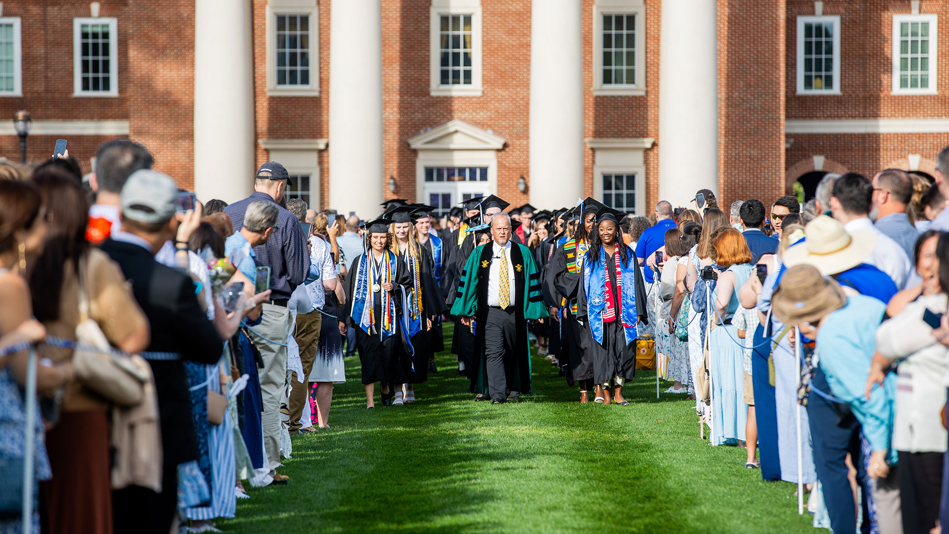- Graduates and faculty in regalia walk down the Great Lawn lined with spectators away from a large brick building with columns in the background.