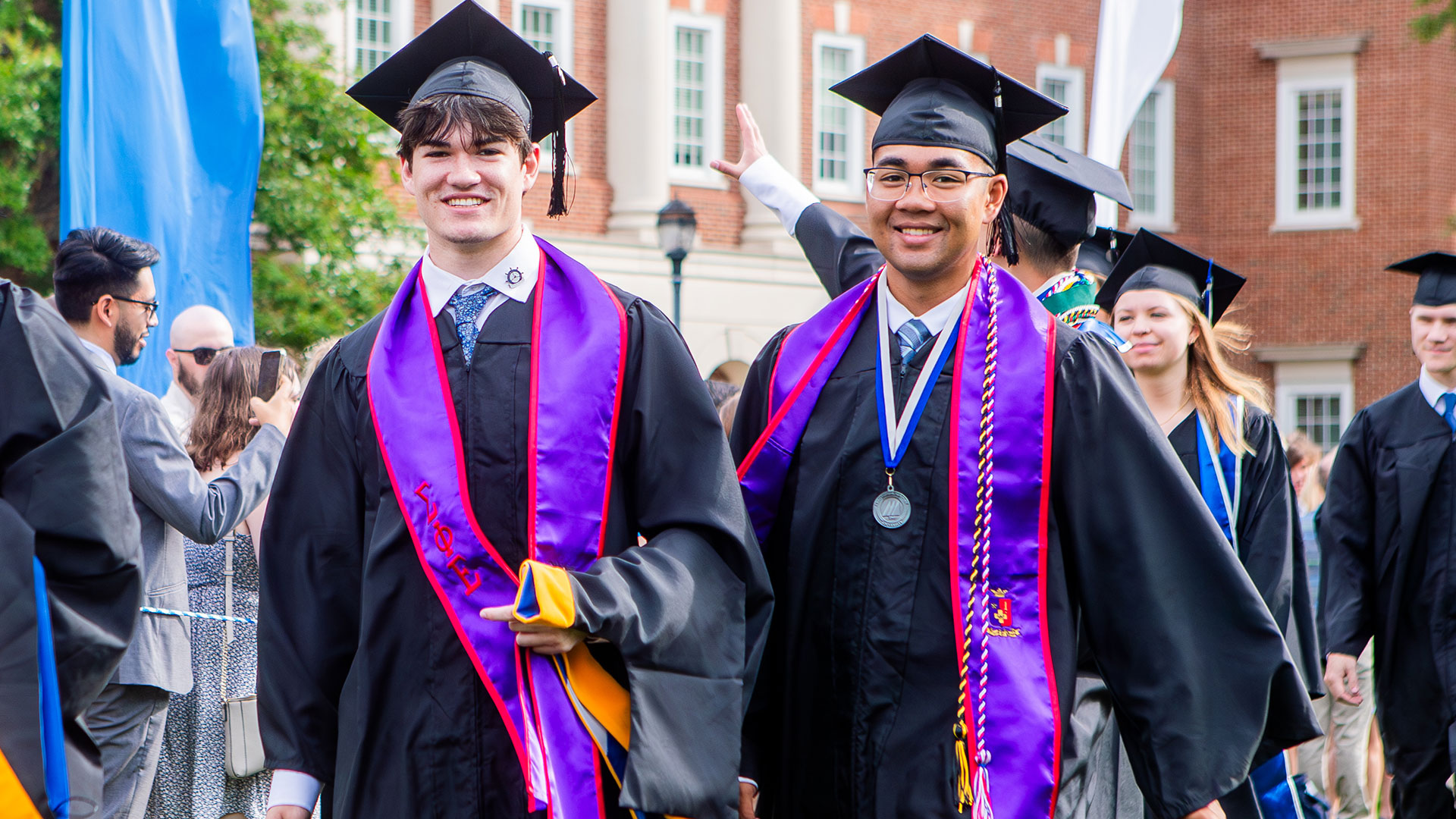 - two smiling graduates in black caps and gowns with purple and red stoles walk toward the camera during a ceremony