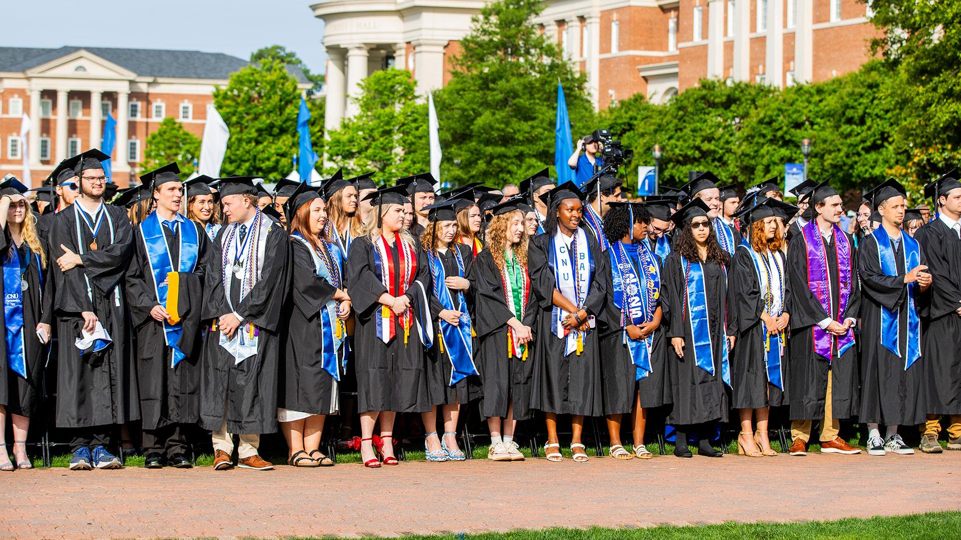 A row of gradutes in black caps and gowns with various colored stoles and cords stand together on the Great Lawn during commencement.