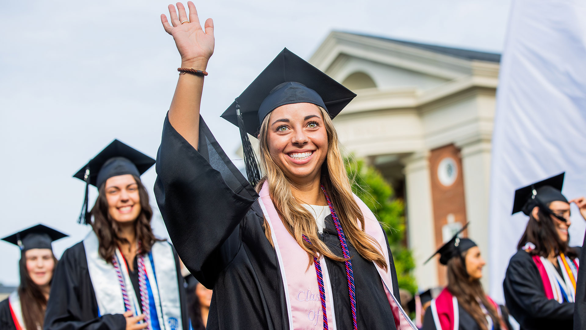 A graduate in a black cap and gown with a light pink stole and purple cord waves to the crowd during the processional.