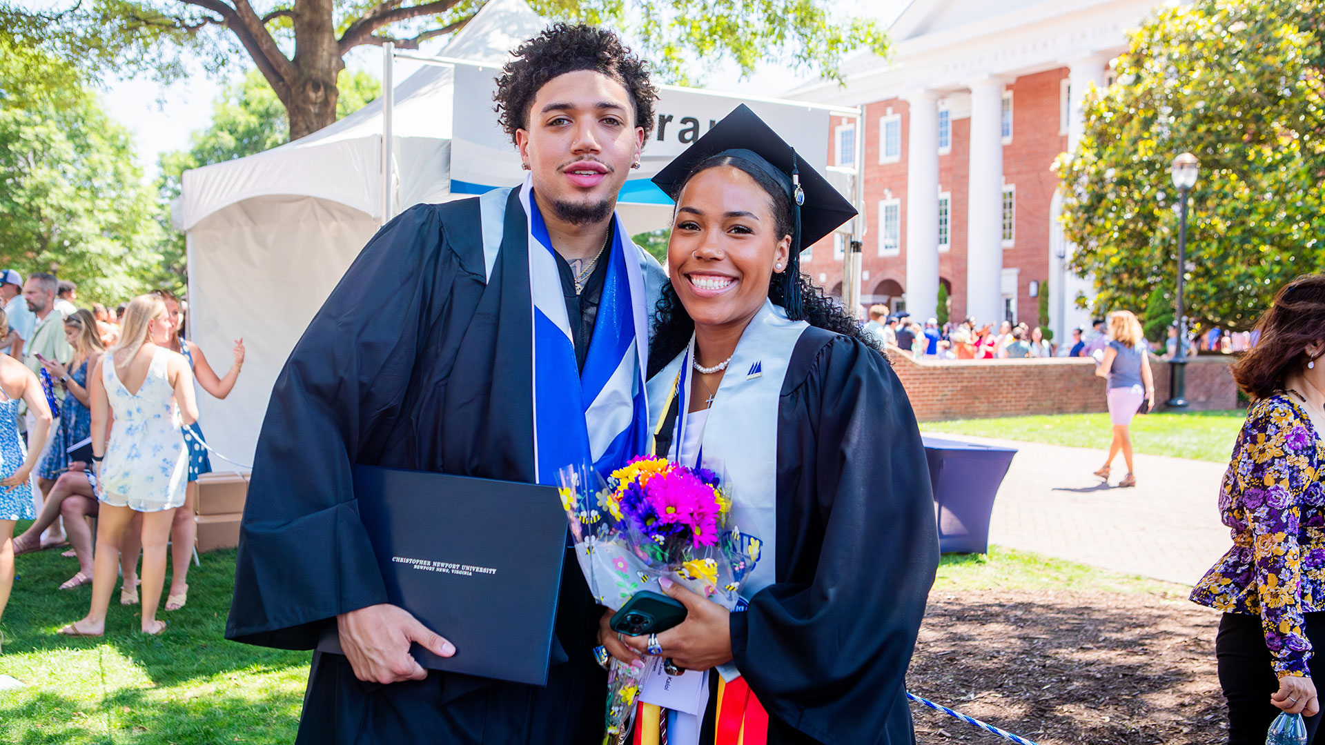 Two graduates standing on the Great Lawn after Commencement. One is holding a bouquet of flowers.
