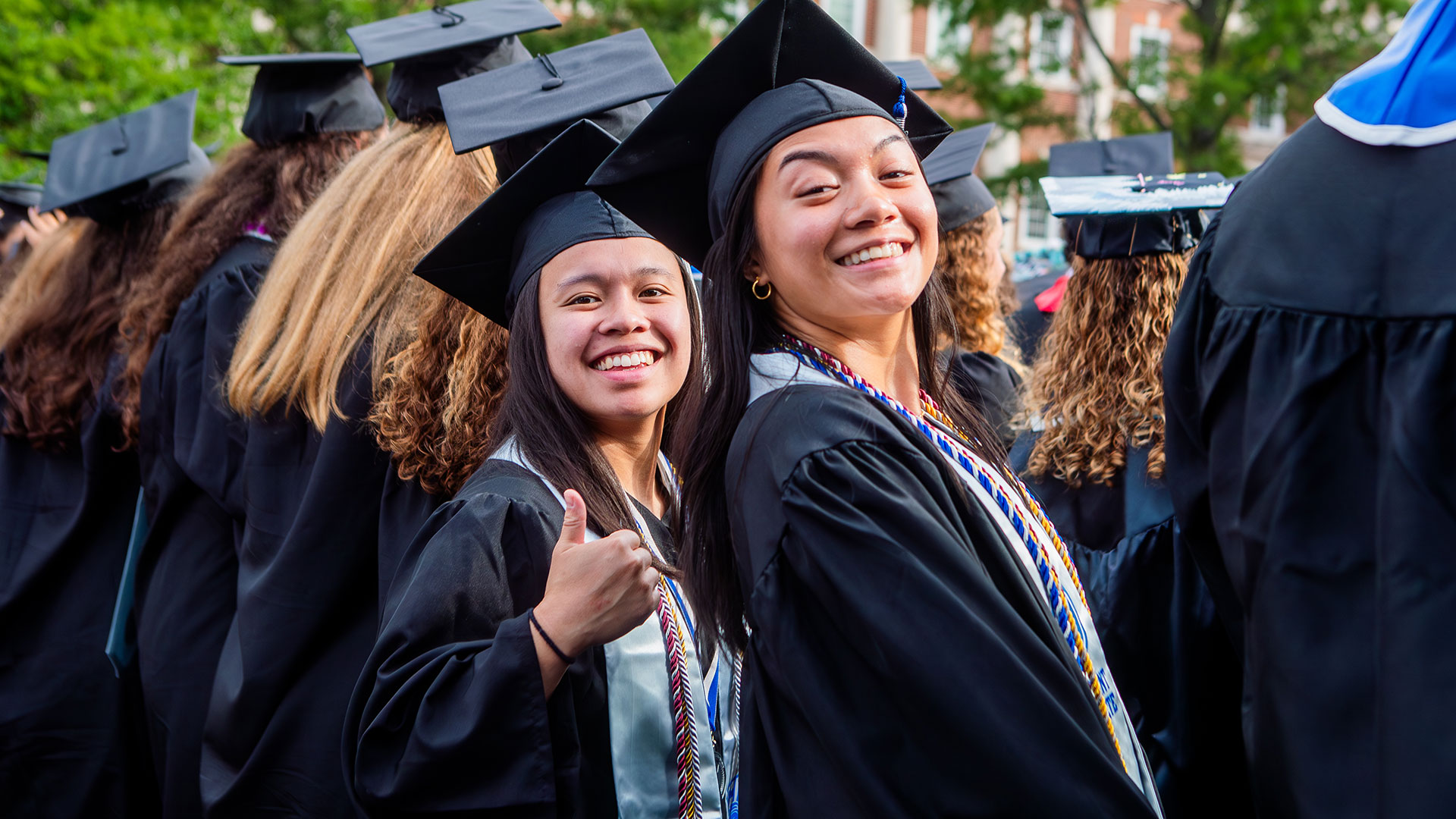 Two graduates in cap and gown smile at the camera and give a thumb's up