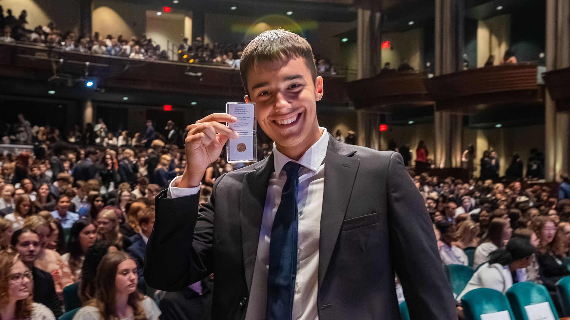 A student wearing a suit holds up the penny given to him after signing the Honor Code.
