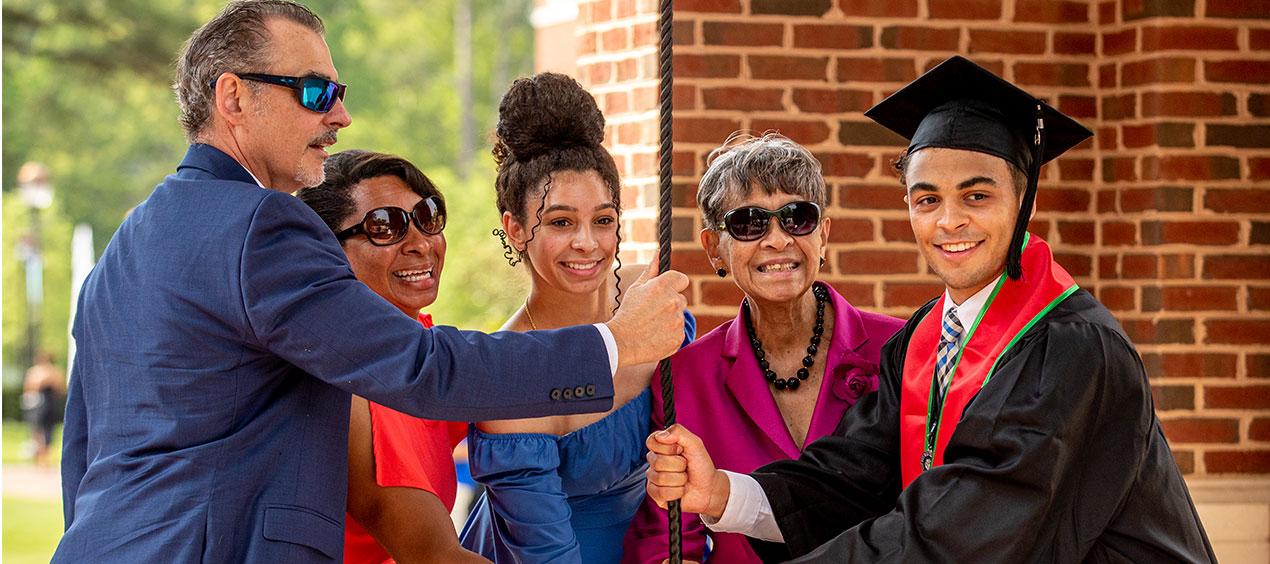 A graduate in regalia ringing the bell tower bell with family