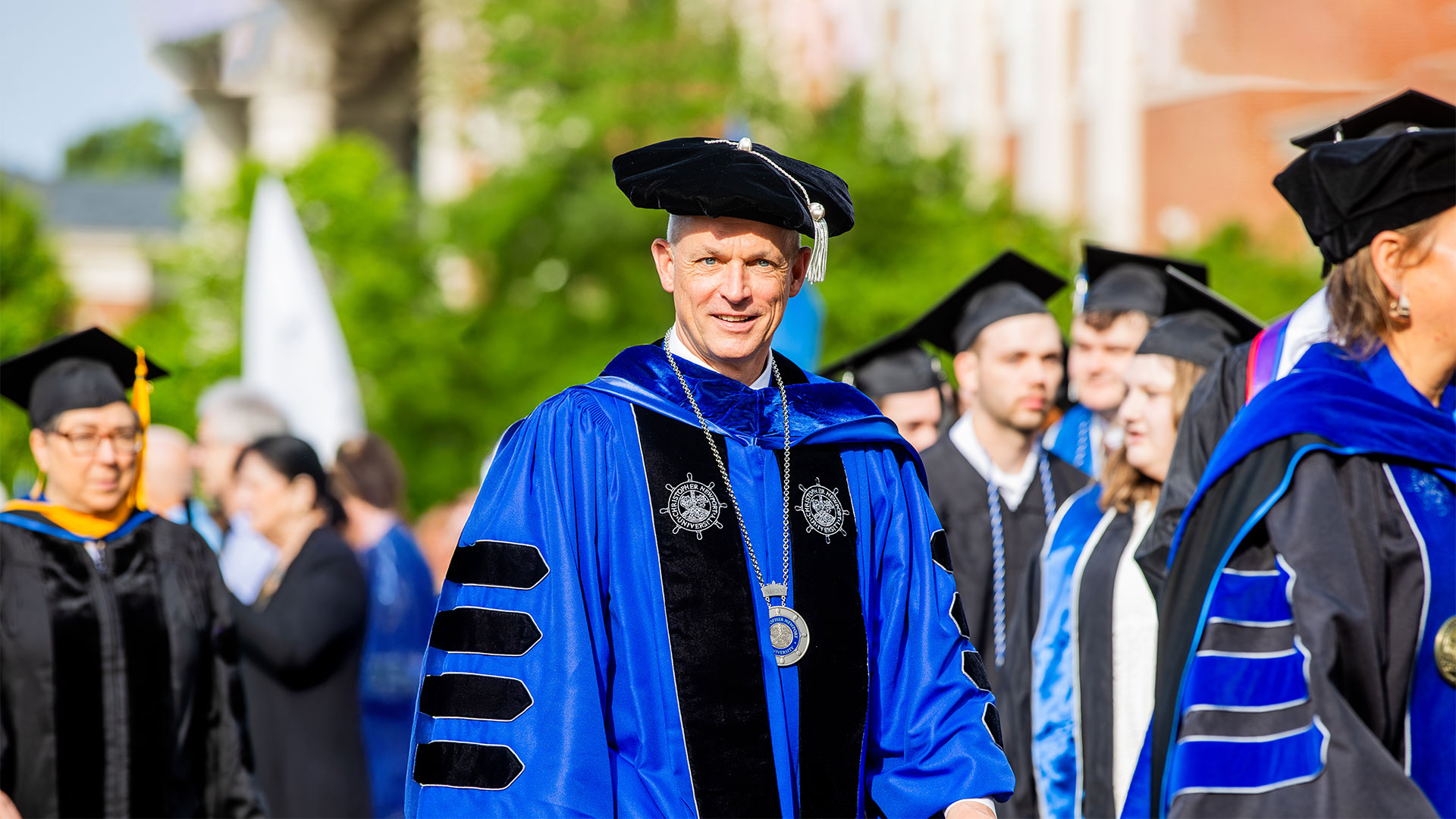 President Bill Kelly in full graduation regalia smiles while walking in the processional