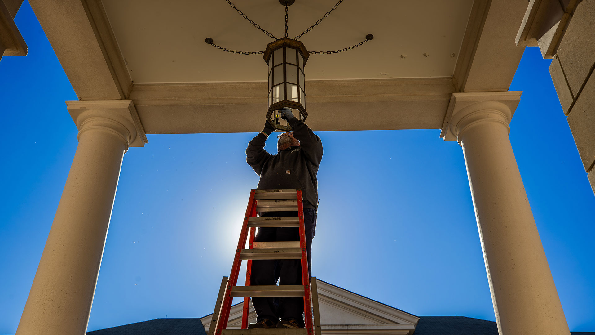  A worker on a ladder replaces a bulb in a large, antique-style hanging lantern under a white portico ceiling against a bright blue sky.
