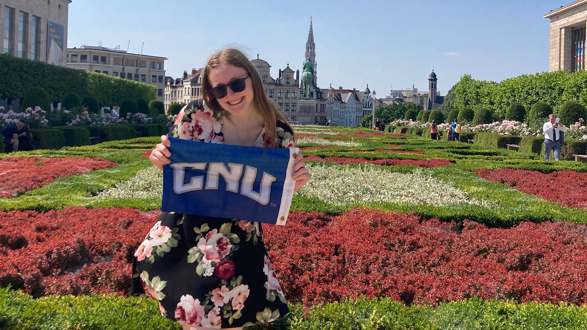A student in a floral dress holding a blue CNU flag in the Mont des Arts garden with the Brussels skyline behind her.