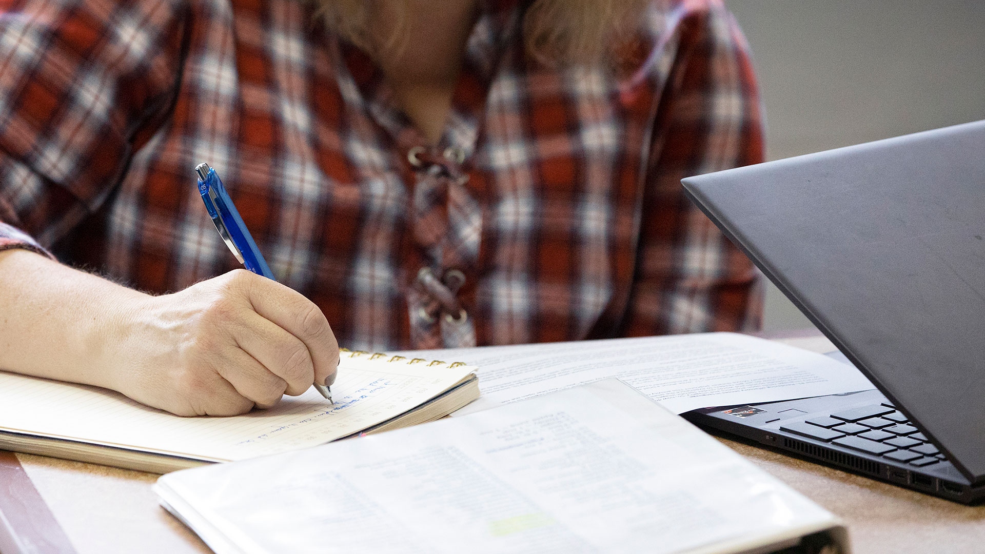 Closeup of a hand writing in a notebook with a binder and laptop on a table