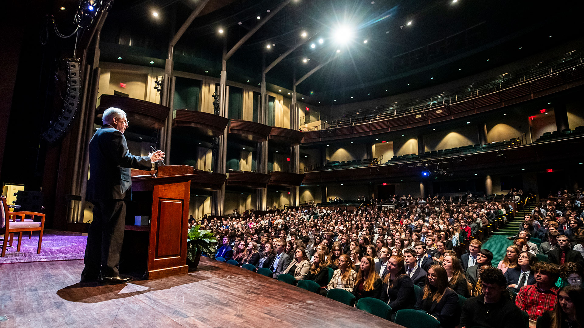 Bob Colvin stands at a podium on stage with a spotlight as students in the President's Leadership Program look on