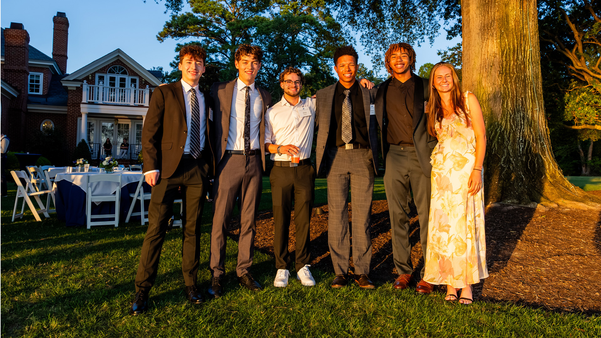 A group of students in business attire gather for a photo during a sunset in the backyard of the university president's house