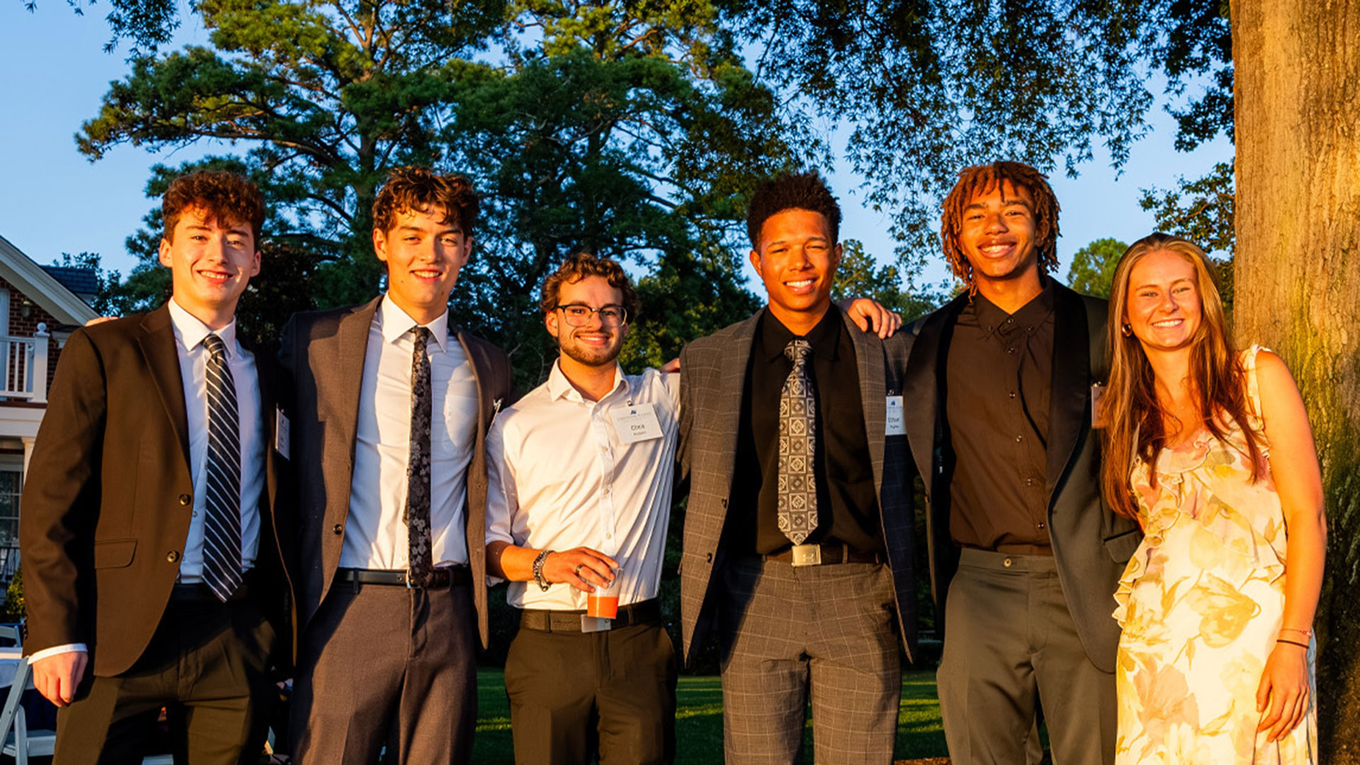A group of individuals in business attire smiling for the camera at sunset while in the university president's backyard.