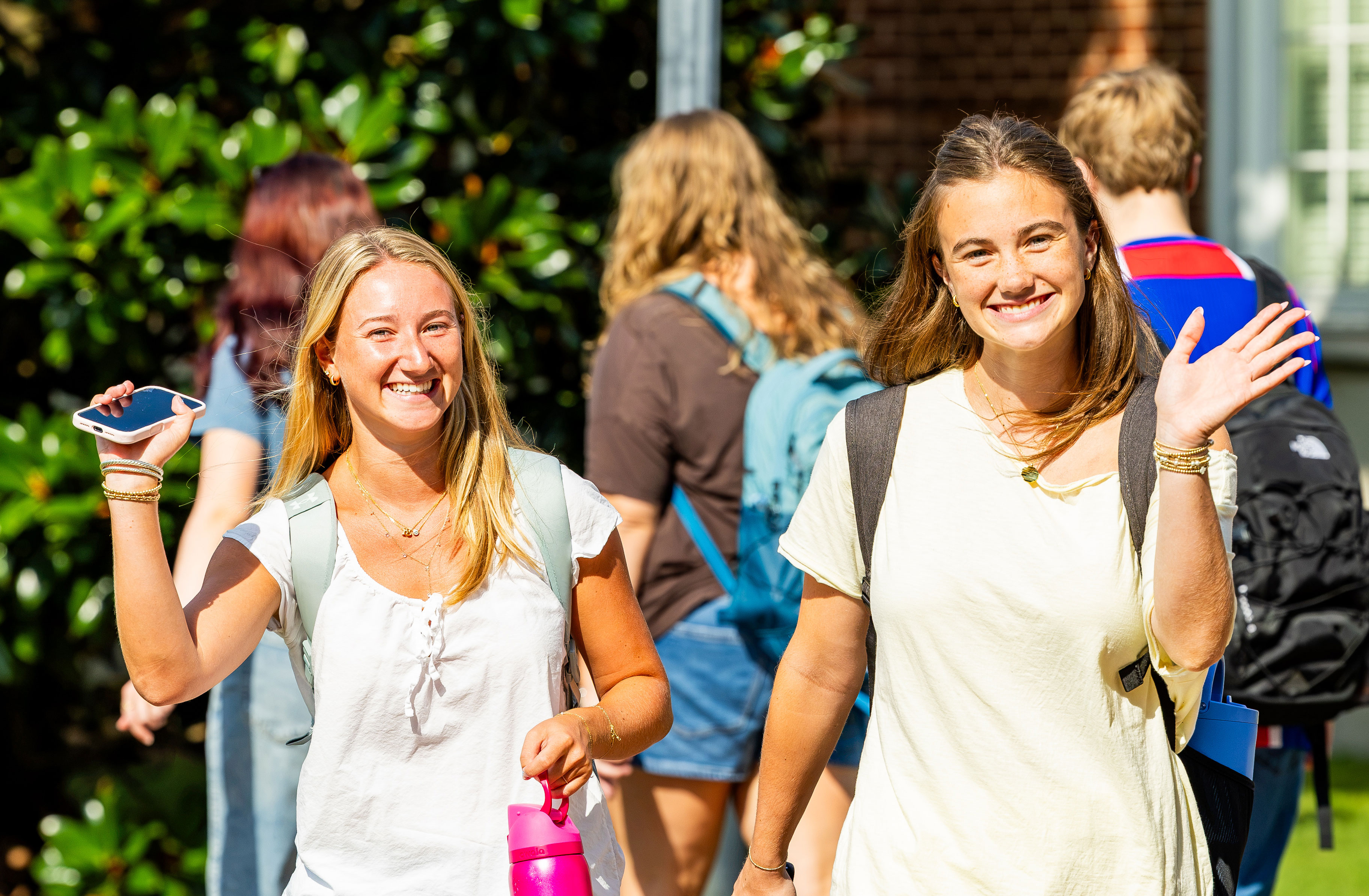 Two women wave to the camera while walking outdoors with backpacks on.