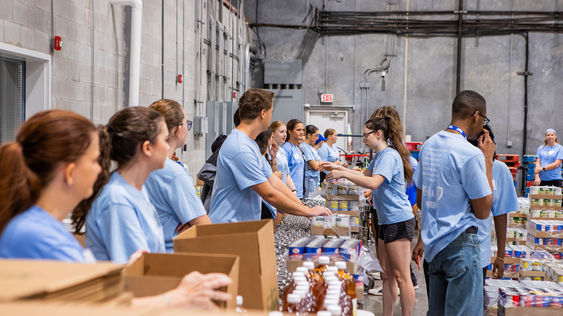 Students in light blue shirts stand on two sides of a conveyor belt line in an industrial warehouse with packaged food in front of them