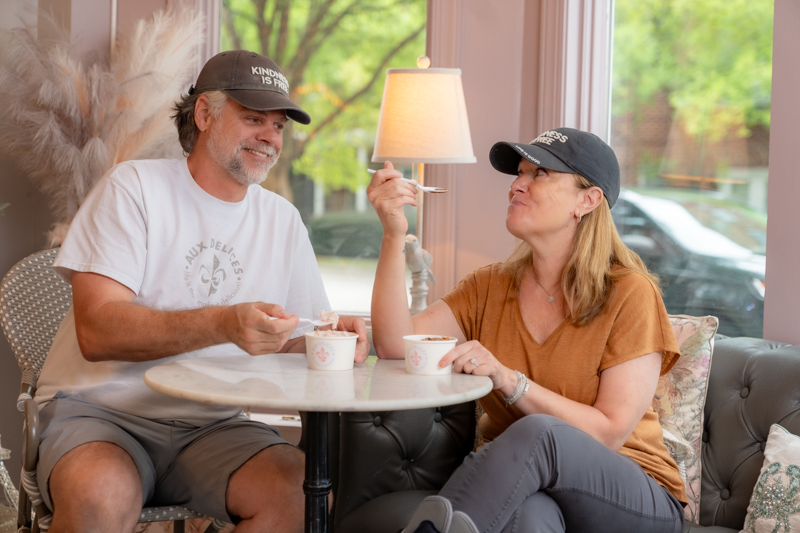 A man and a woman sit eating ice cream, both wearing dark grey hates that say Kindness is Free