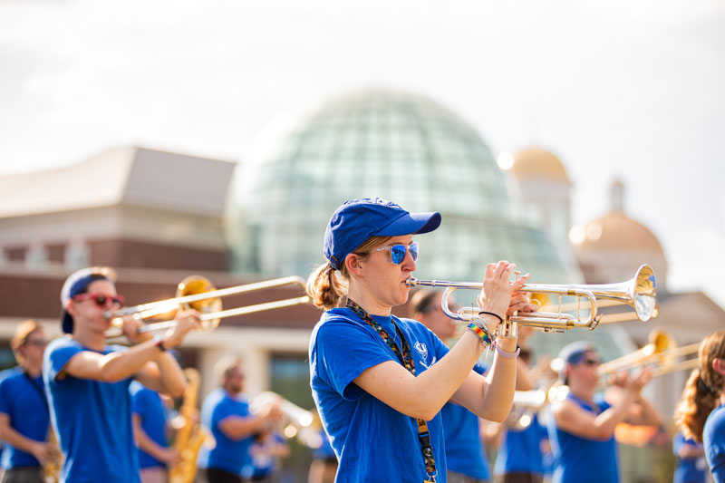 Raechel Erler wear's a hat and sunglasses while playing a trumpet.