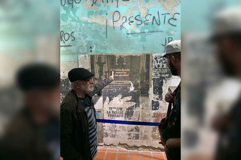 An older gentleman on the right is engaged in conversation with a student on the right. In between them are old posters plastered on a wall.
