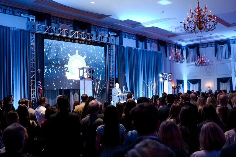 A man is illuminated on stage in the background. In the foreground are students preparing to graduate in the shadows.