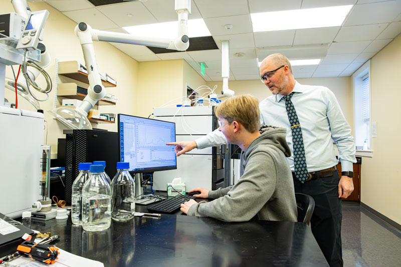 A student sits at a computer in a lab while a professor looks on.