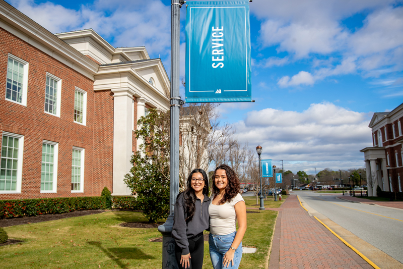 Alexandra Delgado and Niurka Verduzco Rios stand in front of a light post with a light pole banner that reads Service