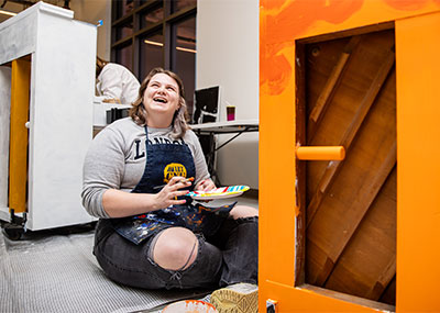 Candyce Harrell smiles while holding a paintbrush in front of her orange piano