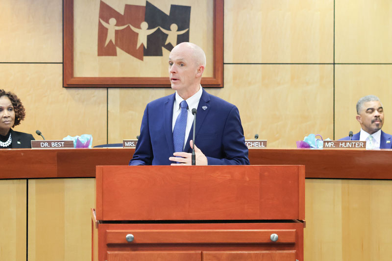 President Kelly stands at a podium facing community members while members of the Newport News School Board look on.