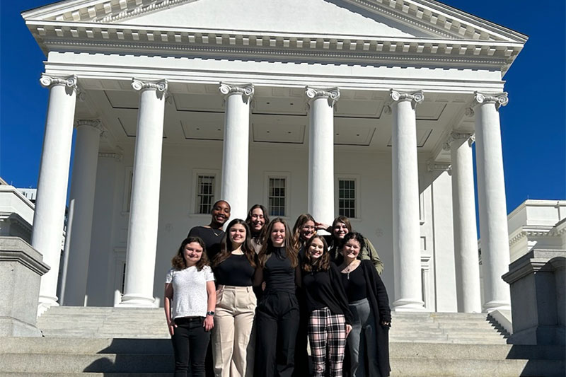 A group of students in dark business professional attire stand on the steps of the capital in Richmond.