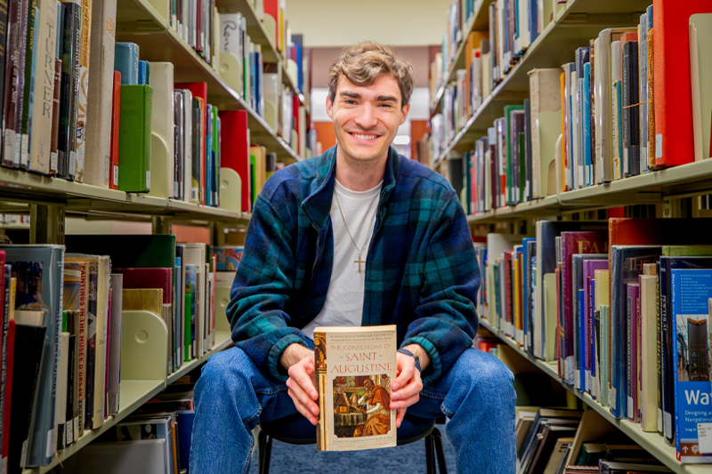 William Slover holds a book while seated between two stacks of library books.