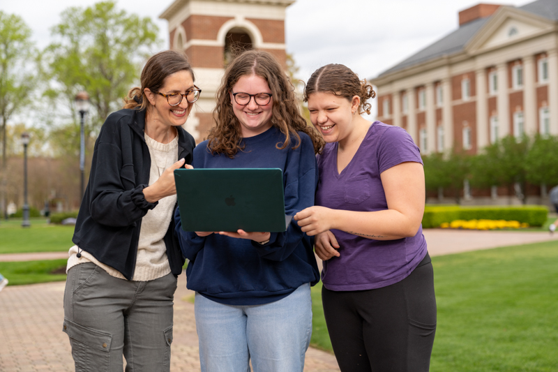 Three women stand and look at a laptop that is being held by the person in the middle.