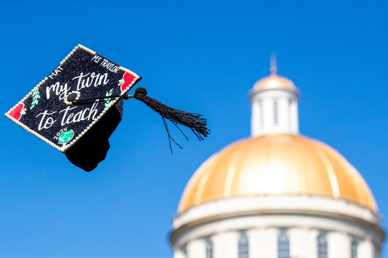 A decorated graduation cap flies in the air with the golden cupola of Christopher Newport Hall out of focus in the background.