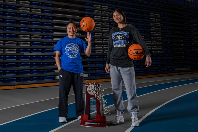 Gabbi San Diego and Camille Malager stand in front of bleachers and behind their national runner-up NCAA trophy.