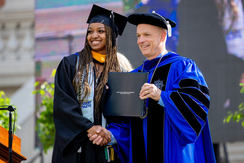 A student shakes hands and holds their diploma with President Kelly.