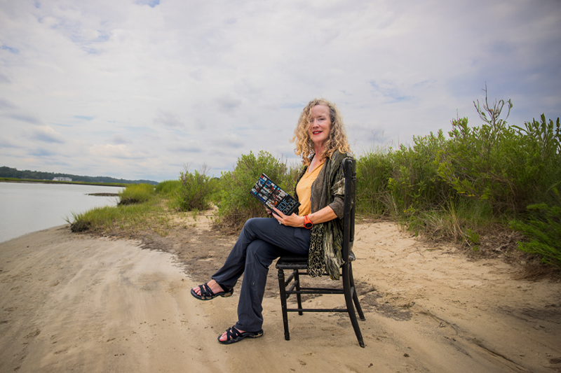 Sharon Rowley sits in a chair on a beach with beach grass blowing behind her.