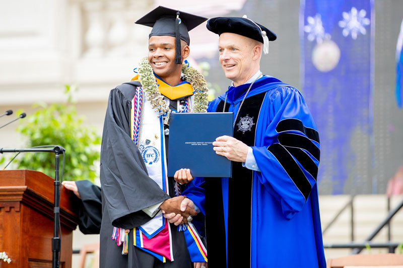 Captain Chris stands besides a graduate who is proudly showing off their diploma.
