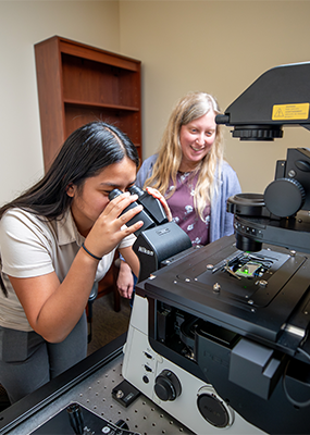 A student looks through a microscope while a professor looks on.