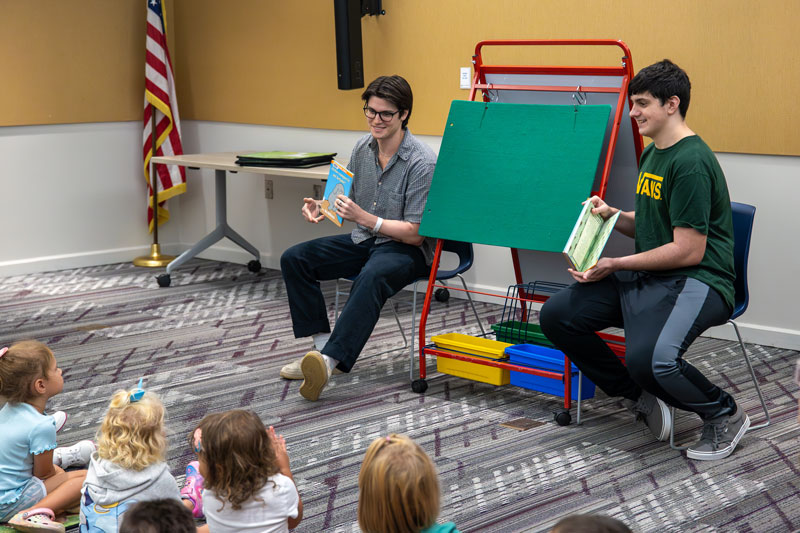 Two students sit in front of a group of young children.