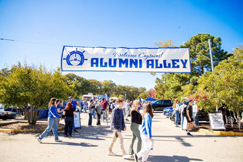 People walk across the entrance to tailgating. A banner that says Alumni Alley hangs over top.