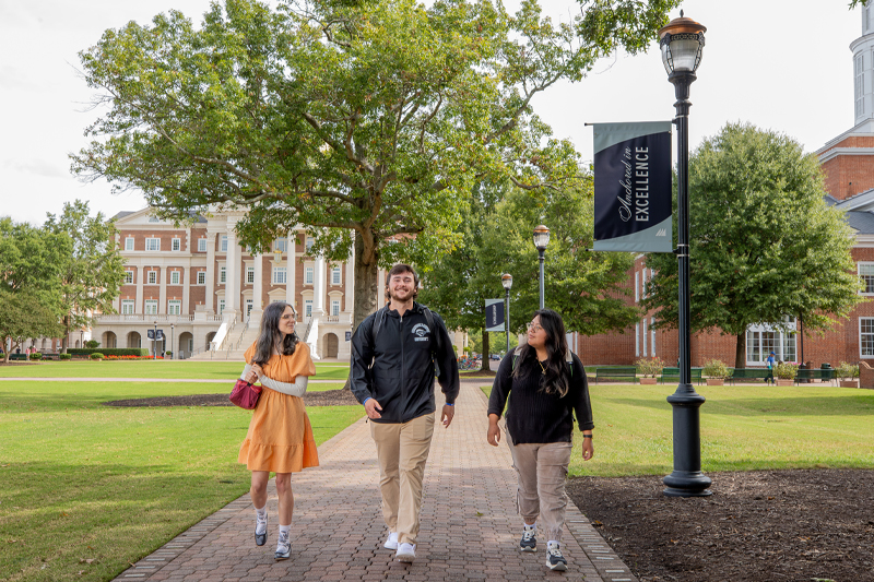Three students walk along a sidewalk next to the Great Lawn with an Anchored in Excellence Banner.