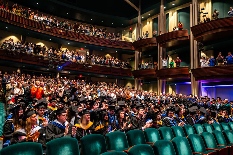 A group of new Christopher Newport graduates are in the foreground of the Diamonstein Concert Hall. Family and friends give them a standing ovation.