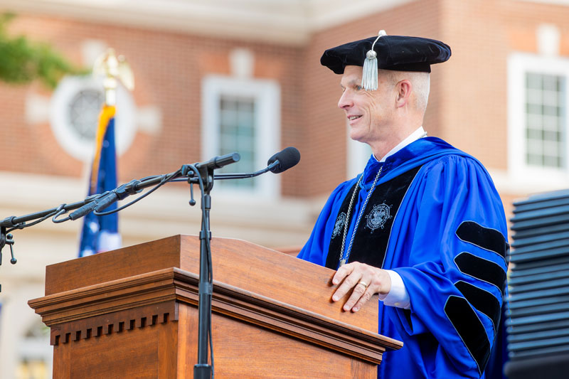 CNU President Kelly stands in graduation regalia at a podium.